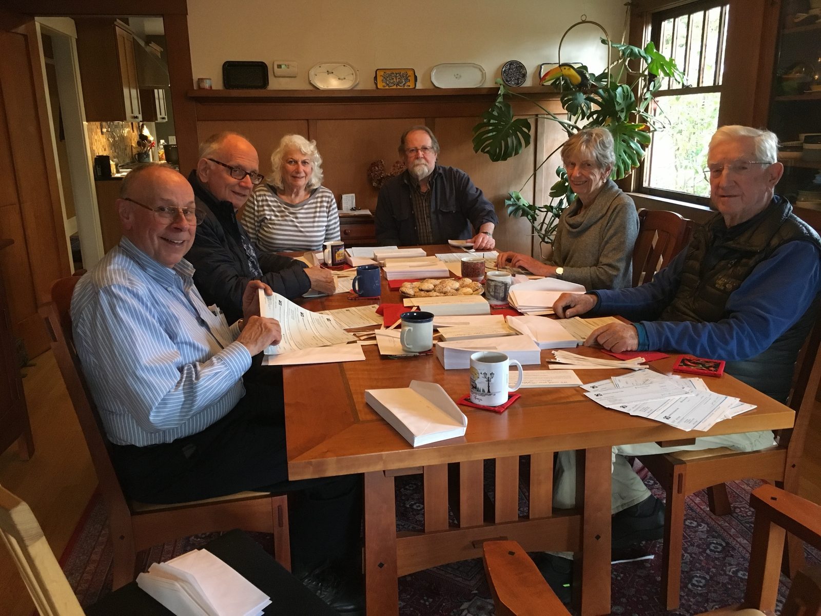 Six board members smiling around a wooden dining table covered in
             stacks of envelopes, coffee mugs and a plate of cookies